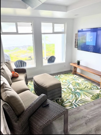 Living room with wicker sectional sofa, tropical leaf rug, large windows with ocean view, and wall-mounted TV